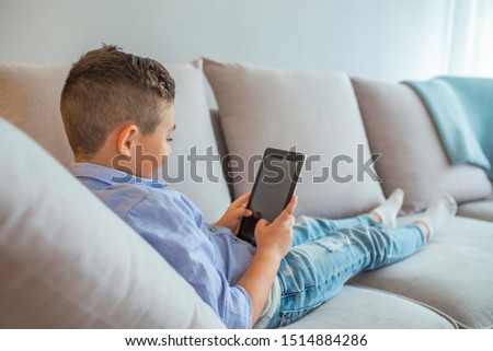 Little boy is smiling while playing with digital tablet at home. Little boy watching cartoons on the digital tablet. Child reads the e-book. Little boy with digital tablet sitting on sofa