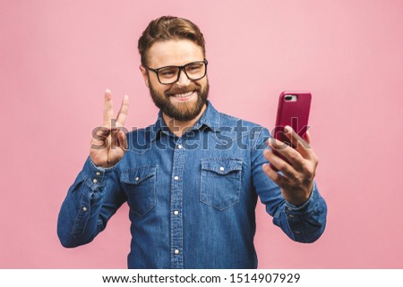 I love selfie! Handsome young man in shirt holding camera and making selfie and smiling while standing against pink background. Ok sign