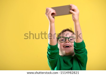 Stylish little boy making selfie. Happy child holding mobile phone. Addicted kid of social networks. Child playing on phone. Kid in green sweater and glasses isolated on yellow background.