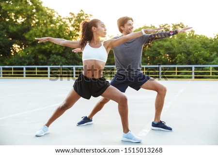 Picture of a concentrated positive young strong sports couple man and woman make yoga exercises outdoors.