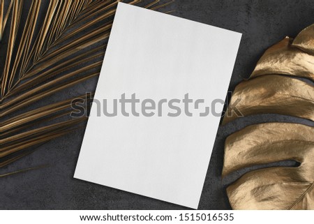 Empty white vertical rectangle poster or card mockup with golden palm and monstera leaves on abstract dark grey textured background. Tropical elegant trendy dark background. Flat lay. Copy space.