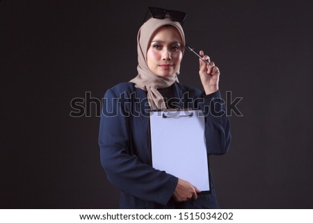 Portrait of a muslim business woman holding pen and blank file while thinking. Isolated on grey background