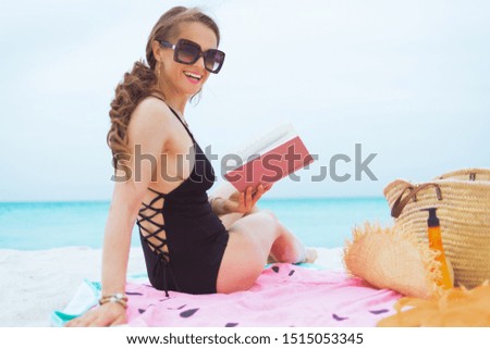 Portrait of smiling trendy middle age woman with long curly hair in elegant black swimsuit with book on a white beach.