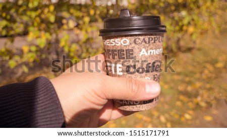Person hand holding a yellow cup of coffee to take away, on an autumn background. 