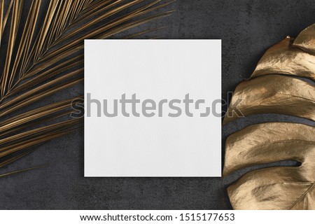 Empty white square poster or card mockup with golden palm and monstera leaves on abstract dark grey textured background. Tropical elegant trendy dark background. Flat lay. Copy space.
