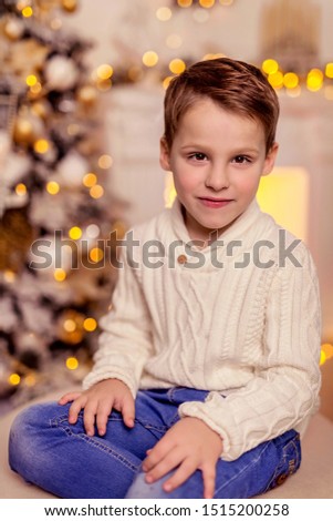 A handsome boy in a white knit sweater sits on an ottoman against the background of a fireplace and a Christmas tree in anticipation of the New Year.