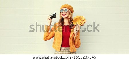 Autumn portrait happy smiling woman holding yellow maple leaves and camera wearing french beret, jacket over gray wall background
