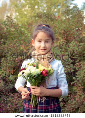 Serious little girl holding flowers and leaves bouquet. Child in school uniform outdoor. Schoolgirl portrait. Girl with thoughtful facial expression. Smiling schoolkid in autumn park