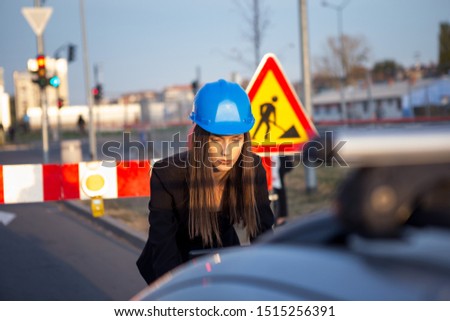 A beautiful young woman construction engineer is on a construction site and she is looking at a laptop located on the hood of a car.