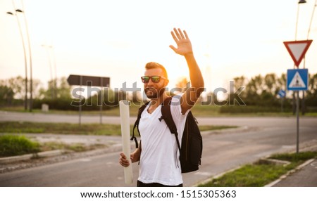 Handsome tourist with sunglasses and a map in his hands. He carries a backpack on his back, stands by the road and waves