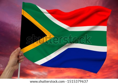 male hand holds the color flag of South Africa on a luxurious texture of satin, silk with waves, closeup, copy space against the sky with clouds