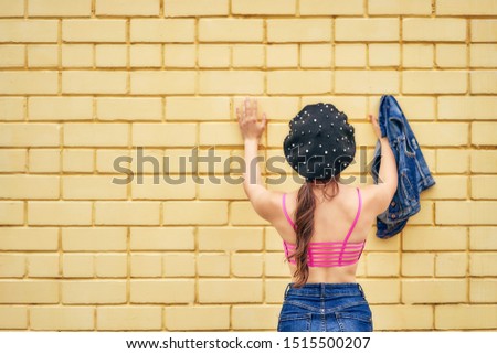 A cheerful woman in a beret and a denim jacket in her hands emotionally poses against a yellow brick wall in a red tank top and jeans. copy space.