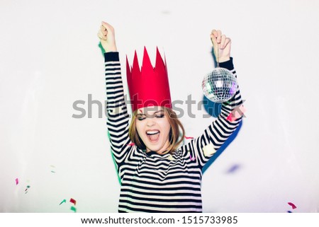 Party girl in colorful spotlights and confetti smile on white background celebrate brightful event, wears stripped dress and red crown. Sparkling confetti, having fun, dancing, laugh, disco ball.