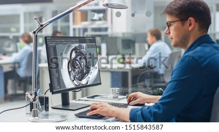 Over the Shoulder Shot of Engineer Working with CAD Software on Desktop Computer, Screen Shows Technical Details and Drawings. In the Background Engineering Facility Specialising on Industrial Design Royalty-Free Stock Photo #1515843587