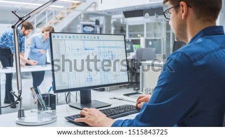 Over the Shoulder Shot of Engineer Working with CAD Software on Desktop Computer, Screen Shows Technical Drafts and Drawings. In the Background Engineering Facility Specialising on Industrial Design Royalty-Free Stock Photo #1515843725