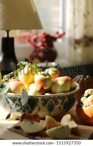Apples in faience fruit bowl, slices, vintage rustic knife on wooden deck, crab apples in pot, pumpkin on tablecloth on kitchen table, vintage style, daylight, natural light and shadow, vertical photo