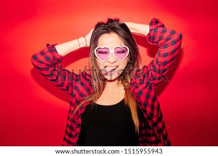Rock'n'roll Kazakh girl in a plaid shirt on a red background playfully shows the tongue in glasses in the shape of hearts, studio portrait