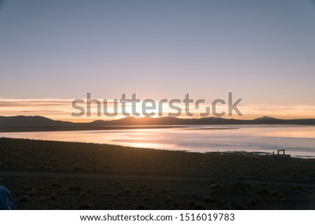 Dramatic view of the sun rising over the lake at sunrise in the morning. View of the lake, sand and rocks. Red, pink, orange, blue sky with clouds. Shot on Salar de Uyuni, Bolivia.