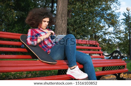 A teenage girl with an african-style haircut sits on a park bench with a skateboard and uses a tablet.