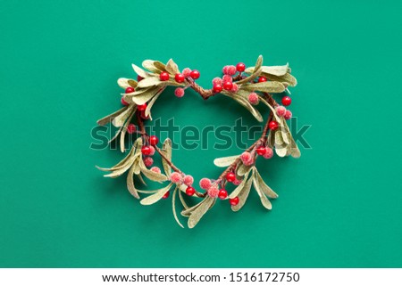 Green paper flat lay with decorative mistletoe with frosted leaves and red berries shaped as a heart