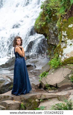 Teen girl in a blue evening dress standing on rocks by a waterfall in late summer