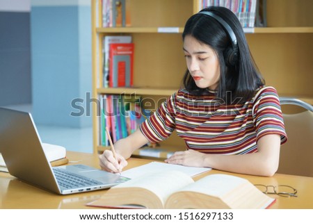 Young Asian woman student study and take notes with book in library. Female university student sits at table does assignments in college library. Student uses laptop and learning online. Education.
