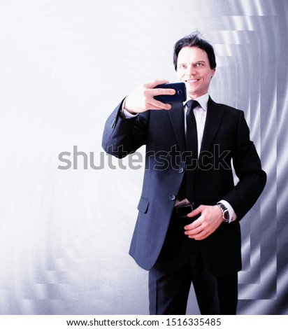 Purposeful adult man in a suit, taking a photo on a smartphone, smiles on a colorful geometric background.