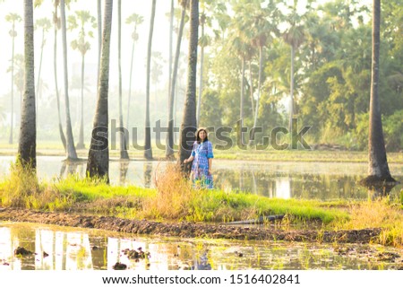 Women wearing a blue dress, wearing a hat standing in paddy field and palmyra palm tree or sugar palm tree in the morning, when the sunrise at countryside of Thailand