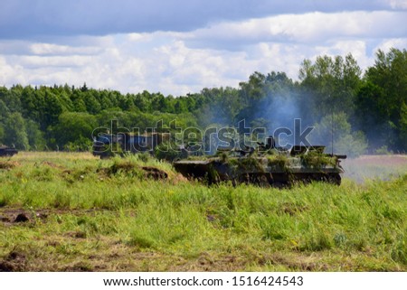 A heavily armoured tank or transporter truck riding along the battlefield generating some smoke with two other military vans in the background seen on a cloudy summer day near a dense forest or moor