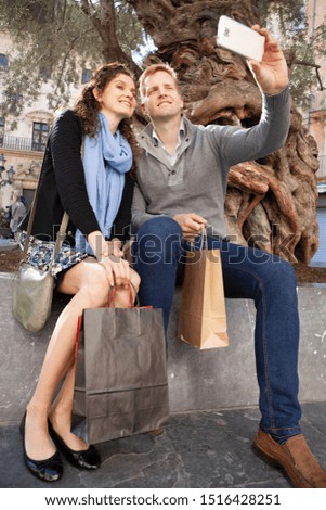 Beautiful consumer tourist couple smiling on sunny city holiday sightseeing with shopping bags, using technology smartphone, taking selfies outdoors. Fun travel vacation, leisure recreation lifestyle.
