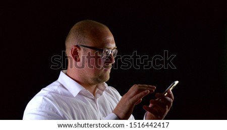 Adult bald bespectacled man holding phone and scrolling screen after typing message, isolated black background