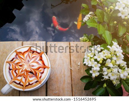 homemade latte art coffee with chocolate sauce and caramel sauce on Milk foam on wooden table beside the fish pond. Fancy carp or koi in the pond and blur flower name andaman satinwood near the table.