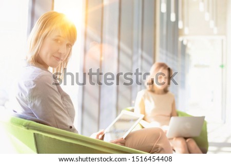 Businesswoman using digital tablet against colleague using laptop in lobby with yellow lens flare