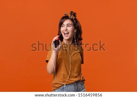Pretty brunette woman in a t-shirt and beautiful headband singing and dancing listening to music with wired headphones isolated over orange background. Enjoying life
