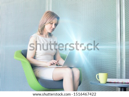 Mature businesswoman working on laptop while sitting and waiting in office lobby with blue lens flare