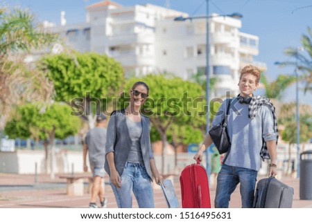 Young smiles looking at the camera as he holds two suitcases and she holds a map in one of his hands and in the background large green buildings and trees. Concept of travel.
