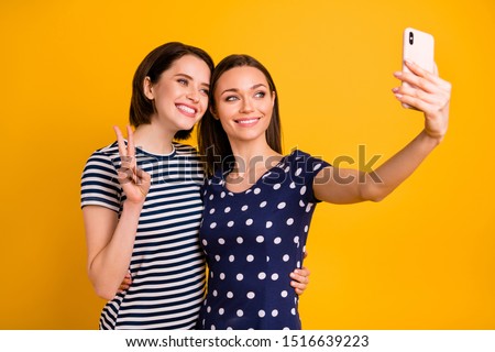Photo of two amazing ladies holding telephone making selfies showing v-sign symbols wear summer casual trendy t-shirts isolated yellow background