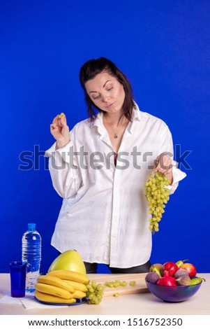 Emotional actress woman in a white shirt posing with fruits in her hands and on the table on a blue background