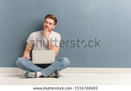 Young redhead student man sitting on the floor doubting and confused. He is holding a laptop. Royalty-Free Stock Photo #1516768682
