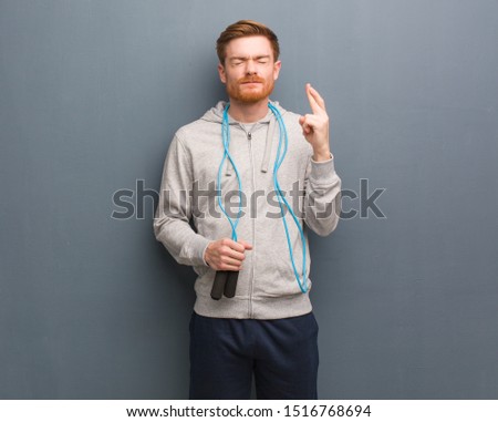 Young redhead fitness man crossing fingers for having luck. He is holding a jump rope.