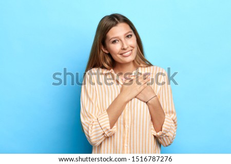 beautiful charming girl keeps both palms on heart, feels gratitude, being touched by serenade, dressed in casual striped shirt, isolated over blue background. Body language concept. happiness, love