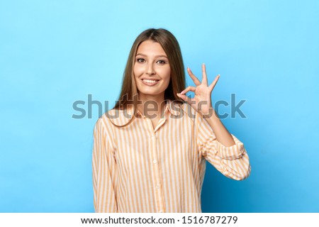 cheerful young beautiful woman in stylish striped shirt shows ok sign with hand as expresses approval, expresses cheerful expression. isolated blue backgroun, studio shot. body language