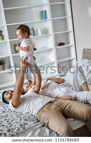cheerful father doing exercises with kid, raising his child up, wjhile lying on bed with his wife . close up side view photo