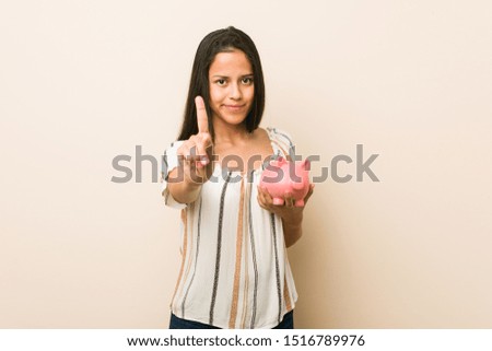 Young hispanic woman holding a piggy bank showing number one with finger.