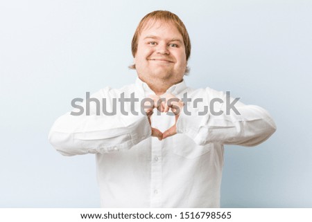 Young authentic redhead fat man smiling and showing a heart shape with hands.