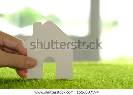 Hand holding white paper house on green grass with blurred office building interior background.