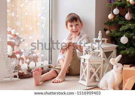 Adorable seven-year-old boy in a cotton white shirt and shorts sits on the floor near a large panoramic window beautifully decorated with Christmas decor. happy child in a cozy New Year's atmosphere