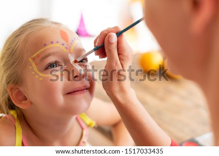 Young mother painting daughters face for Halloween party. Halloween or carnival family lifestyle background. Face painting and dressing up. Royalty-Free Stock Photo #1517033435