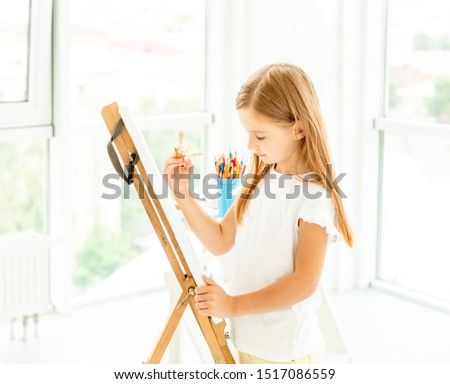 Adorable talented little girl likes to paint different pictures on wide easel