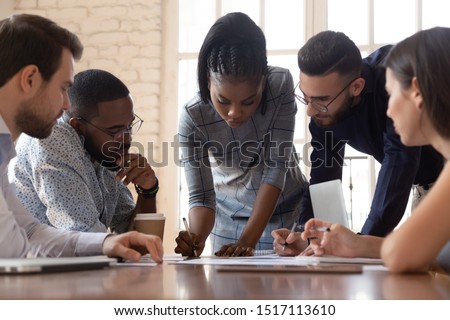 Focused arican american millennial female team leader leaned over table, writing down project ideas, editing documentation at brainstorming business meeting with diverse partners or clients at office. Royalty-Free Stock Photo #1517113610